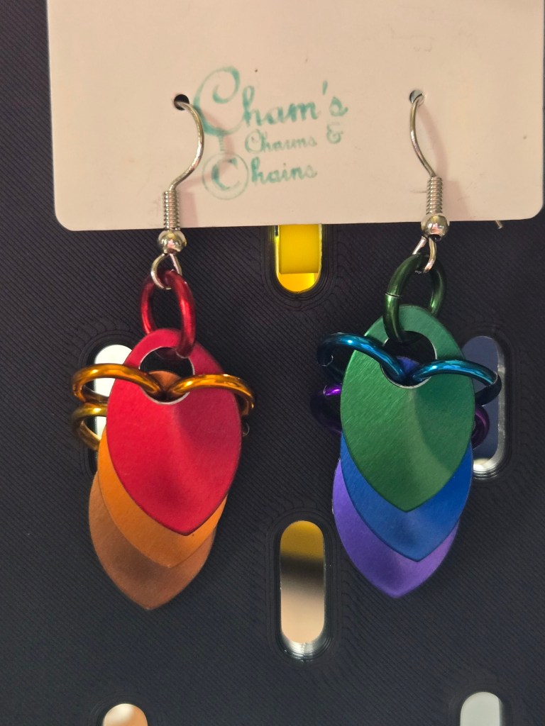 A pair of earrings made of three scales dangling from each other. The six scales are in the colors of the rainbow, red orange and yellow on the left and green blue and purple on the right.