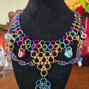 A chainmail necklace of vibrant purple, green and blue rings. A pendant hangs in the center made of rings, with several mushrooms and other beads hanging elsewhere surrounded by twirling chainmail.