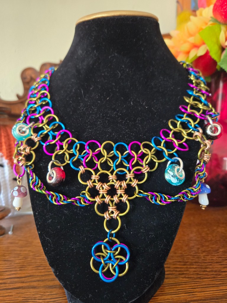 A chainmail necklace of vibrant purple, green and blue rings. A pendant hangs in the center made of rings, with several mushrooms and other beads hanging elsewhere surrounded by twirling chainmail.