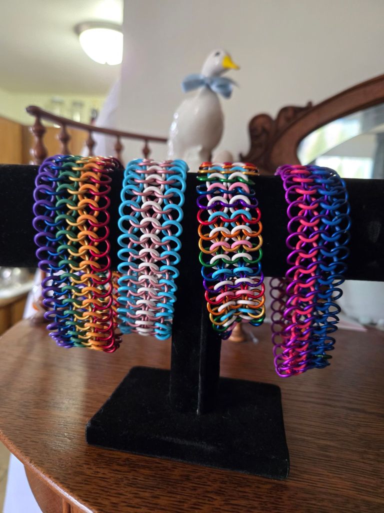 Four thick bracelets on display. They are made of rubber and metal rings in a chainmail patern. From left to right the bracelets are colored as rainbow, transgender flag, rainbow and transgender flag and bisexual flag